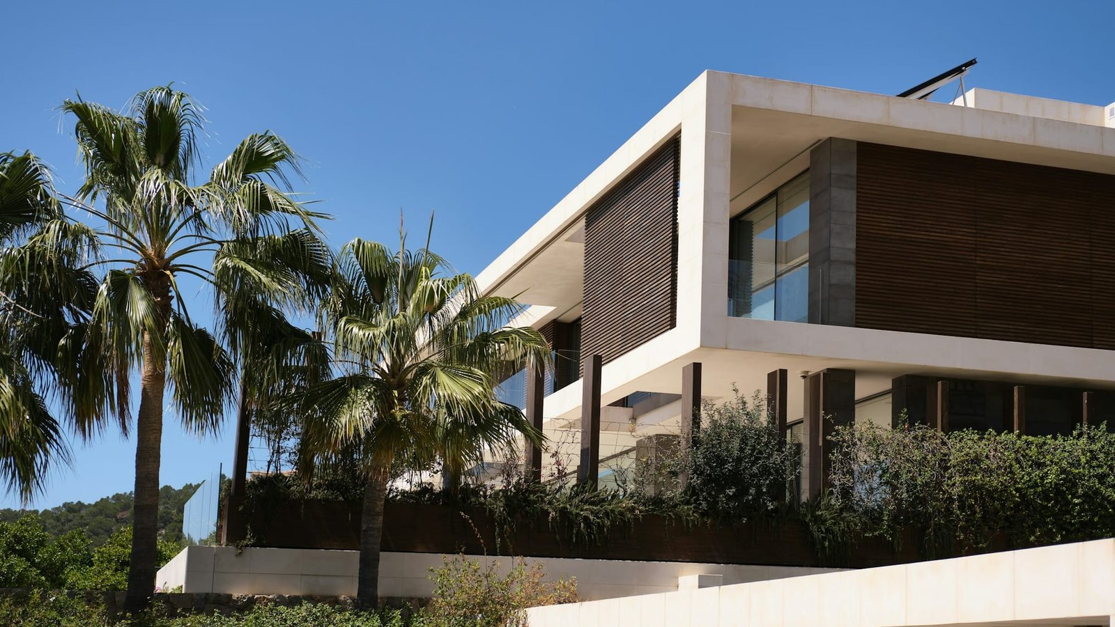 A contemporary stone villa in bright daylight, its upper glazing shaded by deep timber louvres, with palms in the foreground